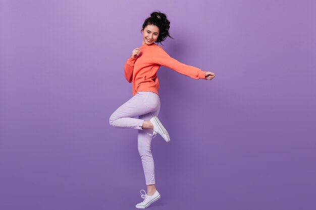 Visão de comprimento total da garota chinesa feliz em pé em uma perna. Foto de estúdio da despreocupada modelo feminino asiático dançando no fundo roxo.