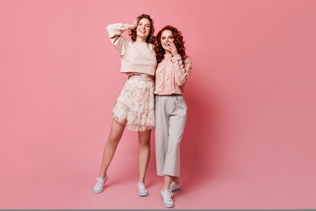 Visão de comprimento total da garota de saia posando com um amigo. Foto de estúdio de duas elegantes jovens em pé sobre fundo rosa.