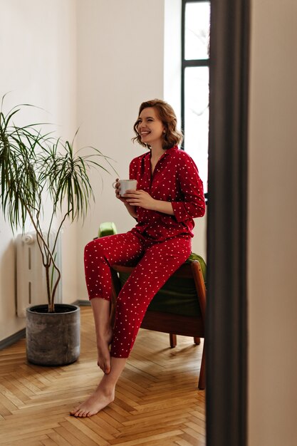 Visão de comprimento total de mulher rindo descalça segurando a xícara de café. Mulher feliz em pijama vermelho, posando de manhã em casa.