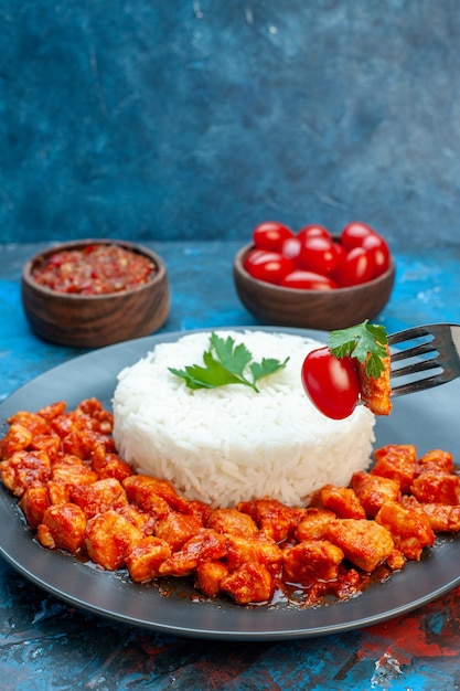 Visão vertical da mão segurando um garfo pegando carne da farinha de arroz com frango e molho em uma placa preta e tomates em uma mesa azul