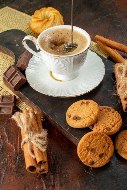 Visão vertical da xícara de café na tábua de madeira, biscoitos, limas e canela, barras de chocolate em fundo escuro