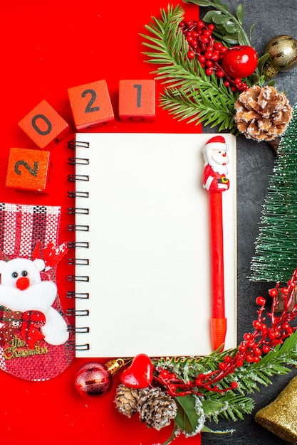 Visão vertical de acessórios de decoração de caderno espiral ramos de abeto xsmas números de meias em um guardanapo vermelho e árvore de natal em fundo escuro