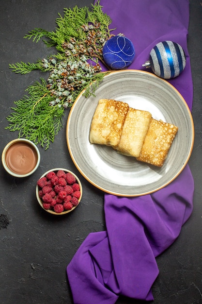 Visão vertical de deliciosos acessórios de decoração de panquecas na toalha roxa e framboesa de chocolate no fundo preto