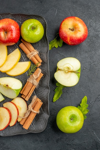 Visão vertical de maçãs verdes fatiadas em fatias vermelhas e inteiras frescas em uma bandeja preta e limão com canela em uma mesa escura