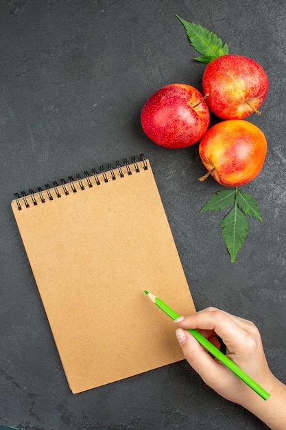 Visão vertical de maçãs vermelhas frescas com folhas e caderno espiral com caneta em fundo preto