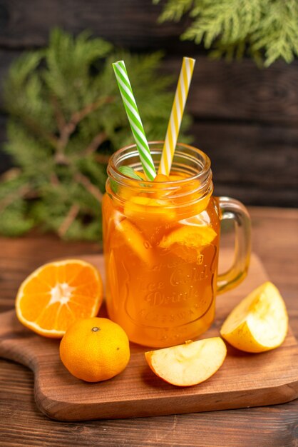 Visão vertical de suco de fruta fresca em um copo servido com tubos, maçã e laranja em uma tábua de madeira sobre uma mesa marrom