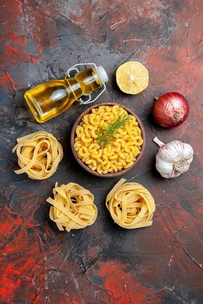 Visão vertical de três spaggeties crus e massas de borboletas em uma tigela marrom e uma garrafa de óleo de alho e limão com cebola verde em um fundo de cor mista