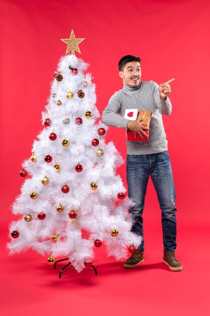 Visão vertical de um homem bonito e sorridente em pé perto da árvore de natal branca e decorada segurando seus presentes