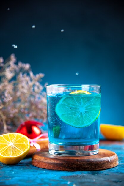 Visão vertical de uma água parada em um copo na tábua de madeira e limão, canela, lima, cone, conífera, acessório de decoração em fundo azul