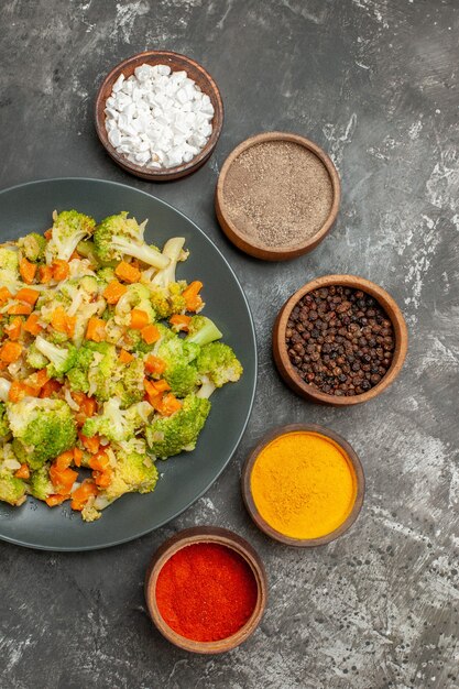 Visão vertical de uma refeição de vegetais com brocoli e cenoura em uma placa preta e especiarias na mesa cinza