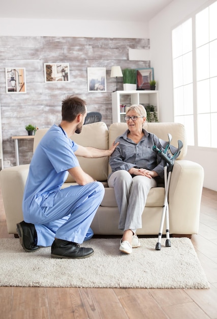 Visitante de saúde masculino em um lar de idosos conversando com uma velha aposentada enquanto está sentado no sofá. Mulher velha com muletas.