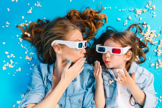 Vista acima da incrível garota alegre deitada com a mãe na pipoca no chão azul. Usando óculos 3D, olhando um para o outro, se divertindo juntos, expressando verdadeiras emoções familiares felizes