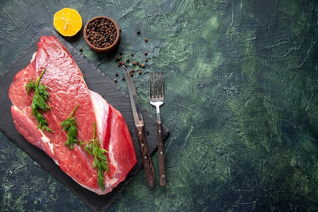 Vista aérea de carne crua vermelha fresca, verde na tábua de cortar e talheres de limão com pimenta no lado direito sobre fundo de cor verde preto