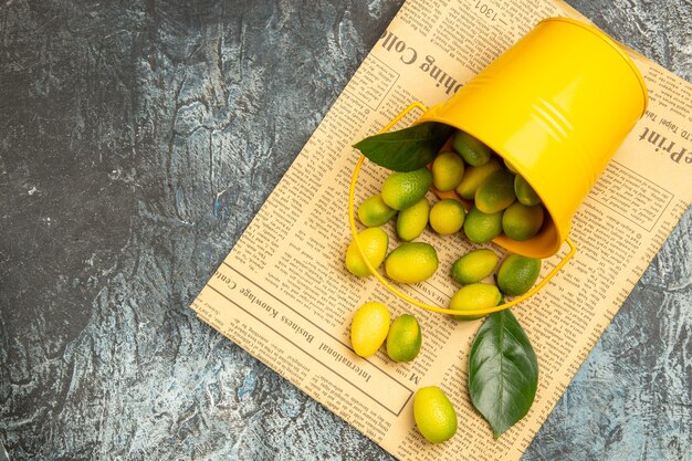 Vista aérea do balde amarelo caído com kumquats frescos nos jornais do lado esquerdo da mesa cinza