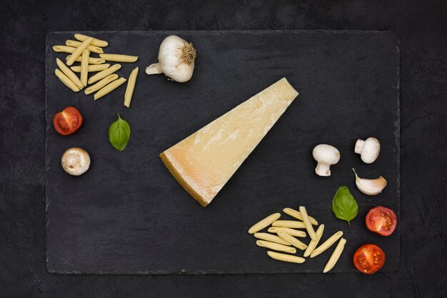 Vista aérea do bloco triangular de queijo com massa garganelli; cogumelo; alho; manjericão e tomate na pedra ardósia sobre o fundo preto