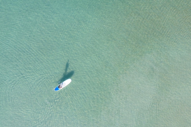 Vista aérea do homem asiático exercitando a prancha de sup em águas claras tropicais turquesa num dia de verão.