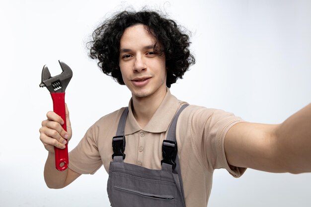 Vista aproximada de um jovem trabalhador da construção civil satisfeito vestindo uniforme segurando chave olhando para câmera isolada em fundo branco