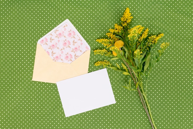 Vista de alto ângulo de flores goldenrod amarelas com cartão; envelope aberto acima de plano de fundo texturizado verde