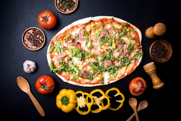 Vista de alto ângulo de pizza saborosa fresca rodeada de legumes; especiarias; e ervas sobre o balcão da cozinha