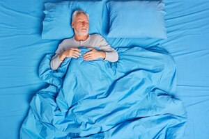 Vista de alto ângulo do homem sênior de cabelos grisalhos barbudo e calmo dorme pacificamente na cama, goza de sonhos agradáveis e sente-se cansado após um dia difícil, vive sozinho posa em um travesseiro azul macio. conceito de madrugada