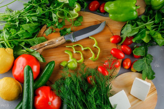 Vista de alto ângulo em fatias de pimenta verde na tábua com tomate, sal, queijo, limão, verduras, faca na superfície cinza