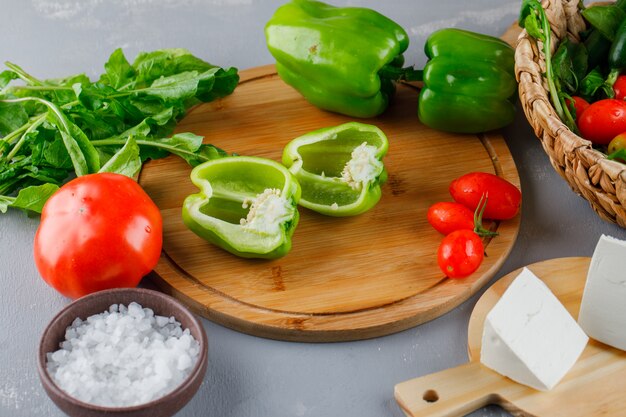 Vista de alto ângulo, pimenta verde cortada ao meio na tábua com tomate, sal, queijo, verduras na superfície cinza