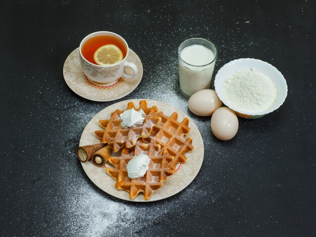 Vista de alto ângulo waffles no prato com chá, ovos, farinha, leite no escuro. horizontal