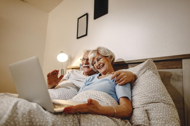 Vista de ângulo baixo do feliz casal sênior relaxando no quarto enquanto faz videochamada no laptop