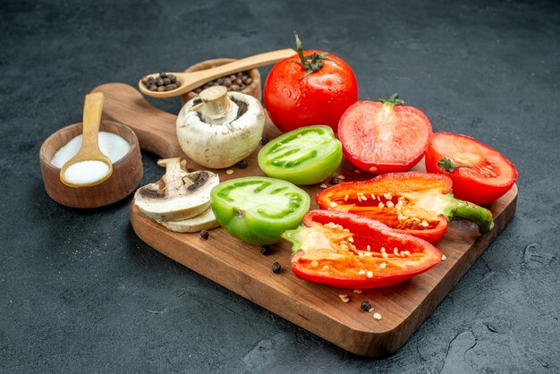 Vista de baixo, cogumelos de legumes frescos cortam tomates vermelhos e verdes pimentões em tigelas de tábua de cortar com pimenta preta e colheres de pau de sal na mesa escura