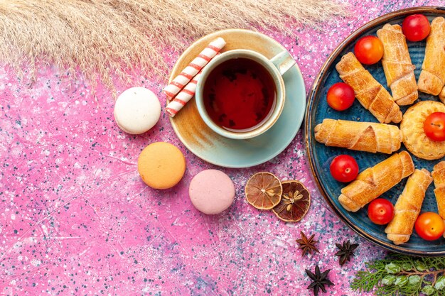 Vista de cima, bagels deliciosos e doces dentro da bandeja com macarons franceses de ameixas azedas frescas e uma xícara de chá na mesa rosa claro
