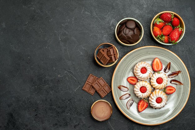 Vista de cima biscoitos doces apetitosos com molho de morango e chocolate ao lado de tigelas de chocolate e molho de morango na mesa escura