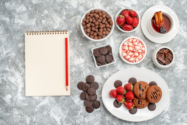 Vista de cima biscoitos morangos e chocolates redondos no prato oval tigelas com doces morangos chocolates cereais uma xícara de chá e lápis de caderno na mesa cinza-esbranquiçada