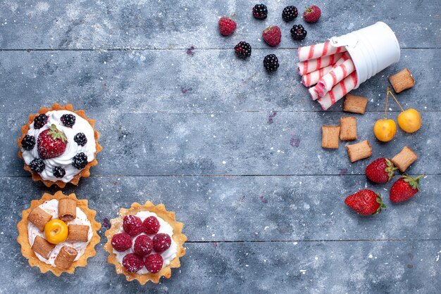 Vista de cima bolinhos cremosos com frutas vermelhas junto com biscoitos de morango e doces em palito rosa na mesa brilhante.
