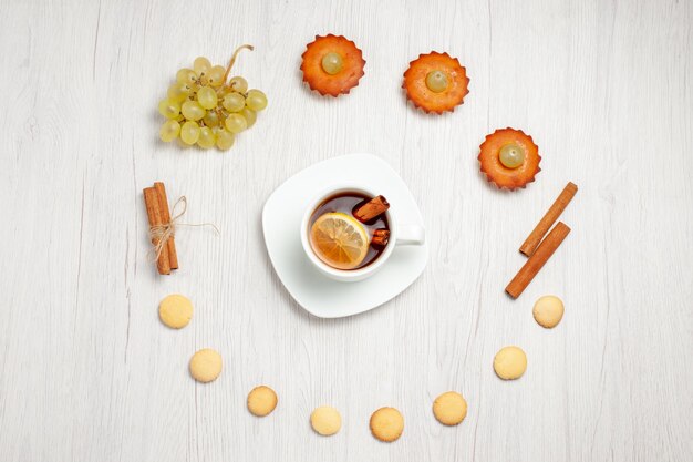 Vista de cima bolinhos saborosos com uvas, xícara de chá e biscoitos na mesa branca, bolo de frutas, biscoito doce, sobremesa, chá