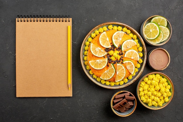 Foto grátis vista de cima bolo com frutas cítricas bolo apetitoso com chocolate e frutas cítricas ao lado do caderno e taças de lápis com fatias de chocolate de limão e creme de chocolate na mesa preta