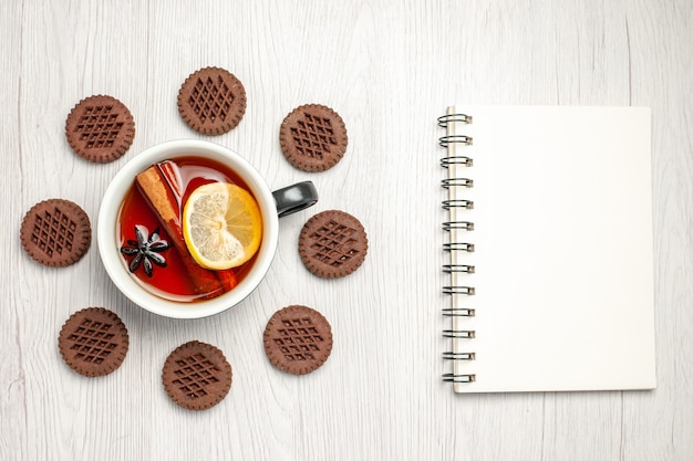 Vista de cima, chá de limão e canela arredondado com biscoitos e um caderno na mesa de madeira branca