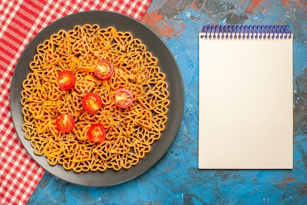 Vista de cima, corações de massa italiana cortam tomates cereja em prato oval no bloco de notas de toalha de mesa quadriculada branca vermelha na mesa azul