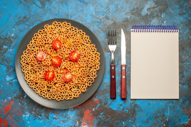 Vista de cima, corações de massa italiana cortam tomates cereja em uma placa oval preta, bloco de notas de garfo e faca na mesa azul