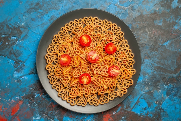 Vista de cima, corações de massa italiana cortam tomates cereja em uma placa oval preta na mesa azul