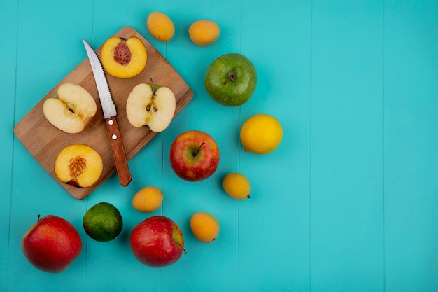 Vista de cima das metades de pêssego com metades de maçã em um quadro negro com uma faca de damascos e lima e limão em uma superfície azul clara
