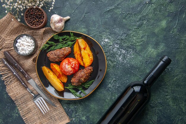 Vista de cima de deliciosas costeletas de carne assadas com batatas e tomates em uma placa preta especiarias talheres de alho conjunto vinho sobre fundo verde preto misturar cores