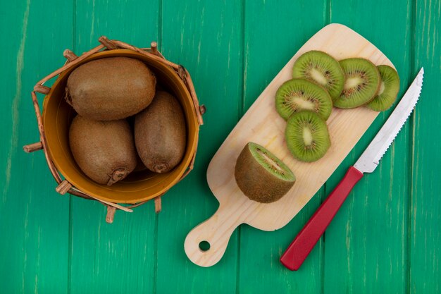 Vista de cima de kiwi em uma cesta com fatias em uma tábua e uma faca em um fundo verde