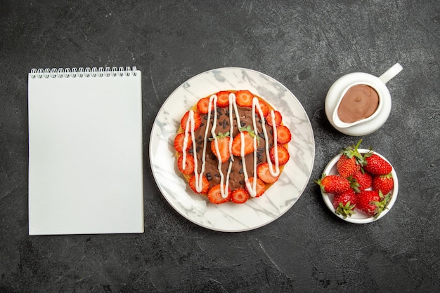 Vista de cima de longe Bolo com bagas Caderno branco taças de morangos e bolo de creme de chocolate com morangos e chocolate na mesa