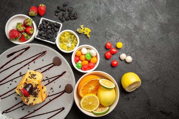 Vista de cima de longe bolo de frutas e frutas com morangos cobertos de chocolate ao lado de tigelas brancas de morangos, limas, limões, laranjas e doces coloridos na mesa