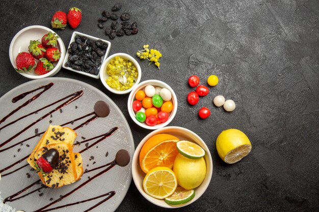 Vista de cima de longe bolo de frutas e frutas com morangos cobertos de chocolate ao lado de tigelas brancas de morangos, limas, limões, laranjas e doces coloridos na mesa