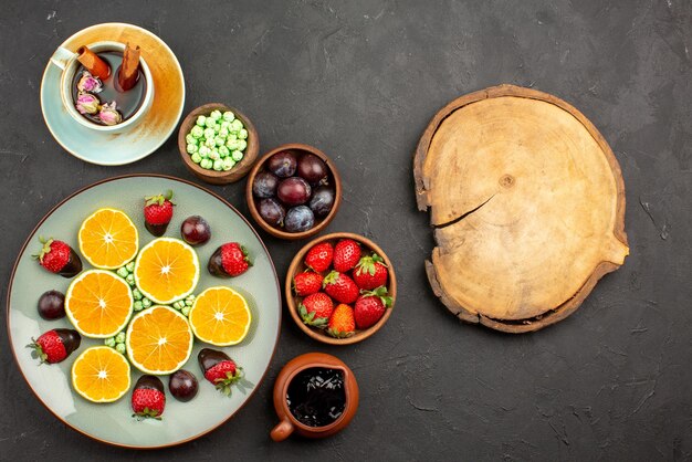 Vista de cima de longe frutas uma xícara de chá uma xícara de chá com cobertura de chocolate, morango picado de laranja e balas verdes e tigelas de frutas vermelhas e doces ao lado da tábua