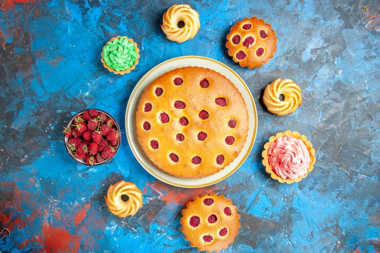 Foto grátis vista de cima de um bolo de framboesa em um prato oval rodeado por biscoitos de uma tigela de tortinhas com framboesas na superfície azul