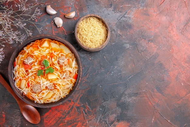 Vista de cima de uma deliciosa sopa de macarrão com frango e macarrão cru em uma pequena tigela marrom e uma colher de alho no fundo escuro