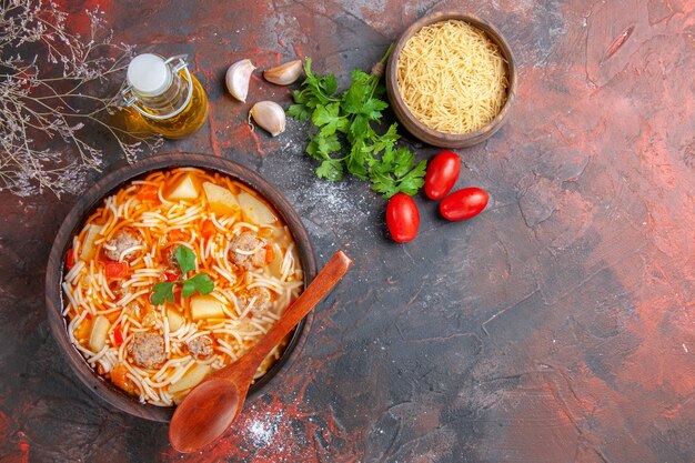 Vista de cima de uma deliciosa sopa de macarrão com frango e macarrão cru em uma tigela marrom pequena e colher tomates de alho e uma garrafa de óleo de verduras no fundo escuro