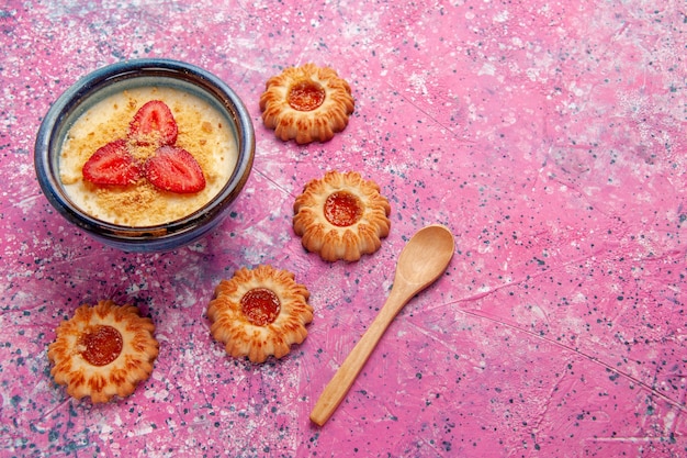 Vista de cima deliciosa sobremesa cremosa com morangos fatiados em fatias vermelhas e biscoitos no fundo rosa.